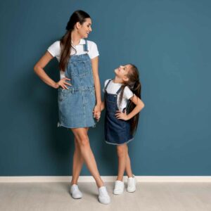 Mor og datter iført matchende denimkjoler og hvide sneakers, smilende og holdende i hånd foran en blå væg – et stilfuldt og hyggeligt look til mor og datter outfits.